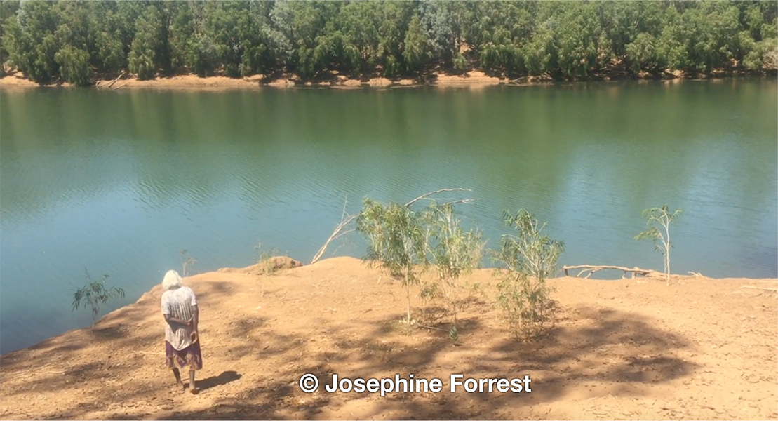 Elderly Aboriginal woman walks down towards the banks of the Martuwarra (Fitzroy River) on a sandy bank with more vegetation visible on the other side of approximately 50m of water.