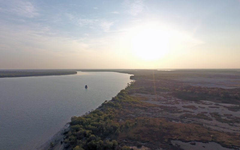 Mitchell River drone image, looking down on mangroves and floodplains to the right and river winding away to the left with a research boat as a small dot in the distance.
